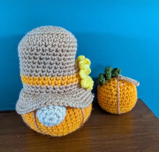 Barnaby, a crocheted harvest gonk in a grey brimmed hat and mustard body, beside a small orange pumpkin