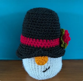 Percy, a crocheted snowman gonk in a black top hat with a red band and an orange carrot nose