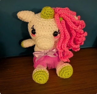 Rosie, a cream crocheted baby unicorn with a coral mane, gold horn and a pink bow