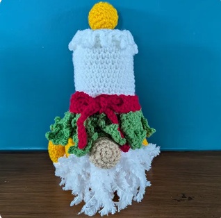 Wick, a white crocheted candle gonk with a yellow flame and a collar of holly and berries